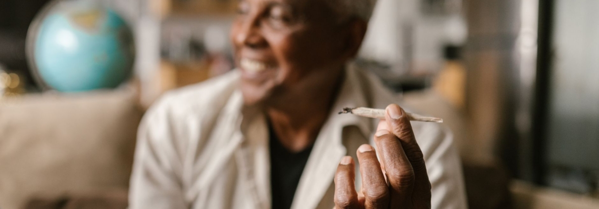 A senior man holding a cannabis joint, relaxing indoors with a blurred background.