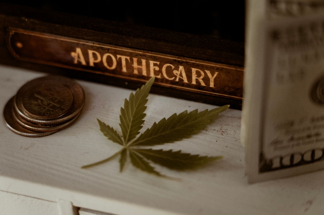 Close-up of a vintage apothecary book, coins, and a cannabis leaf on a wooden surface.