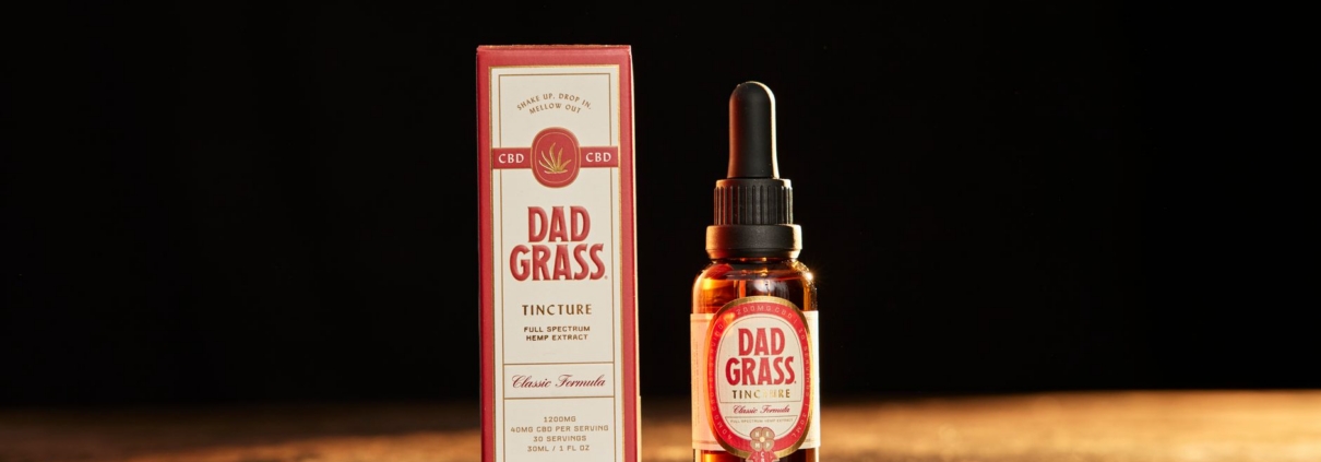 Close-up of Dad Grass full spectrum CBD tincture bottle and box on a warm wooden surface.