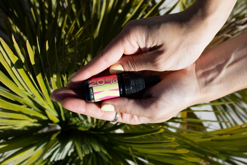 Close-up of hands holding a CBD oil dropper bottle over green palm leaves.