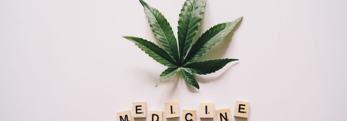 Flat lay of cannabis leaf and 'MEDICINE' spelled with Scrabble tiles on white background.