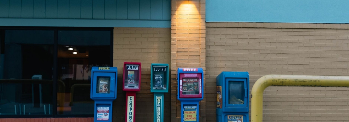 Row of colorful newspaper stands against a brick wall in urban setting during daytime.